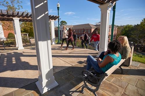 Two students on a bench in the foreground chat as other students crest the stairs to the patio outside the college library.